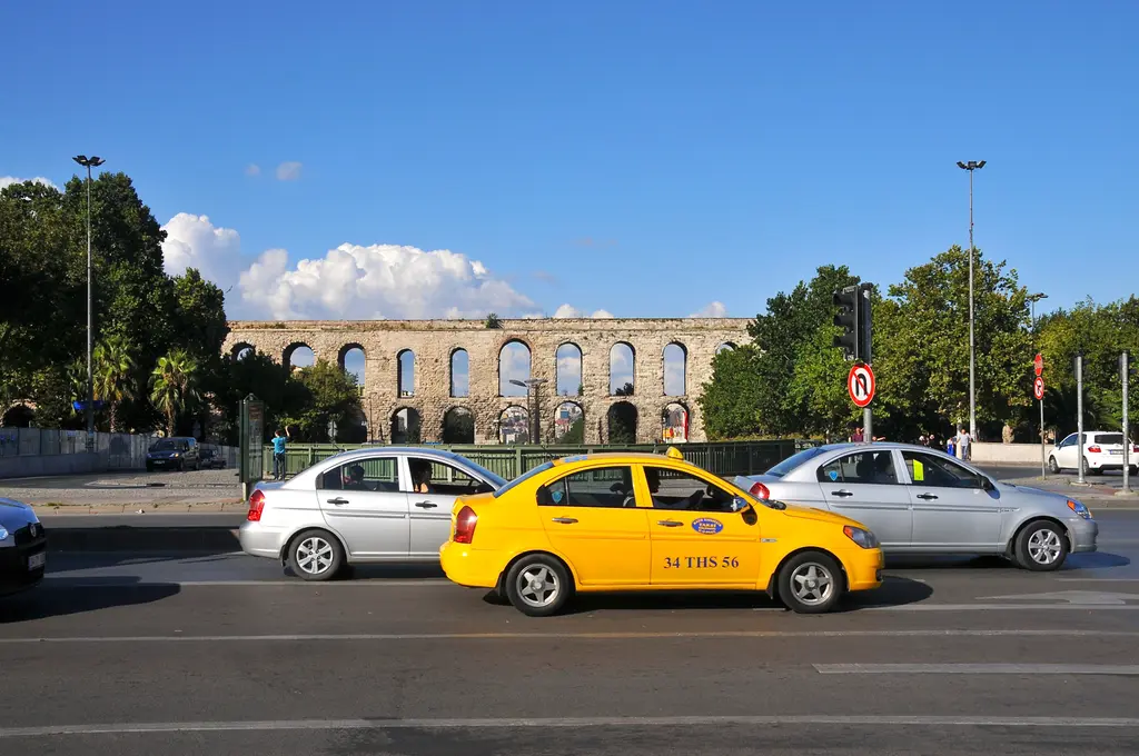 Bozdoğan Kemeri berrak mavi gökyüzü altında; ortada yatay bir bant gibi duruyor, önde geniş asfalt kavşak, sarı taksi, gümüş otomobiller, palmiye ve yeşil ağaçlar.