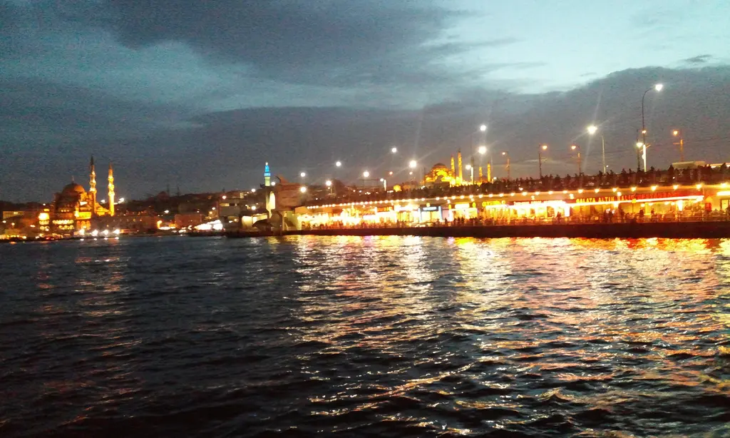 Karaköy'den gece Galata Köprüsü; köprünün ışıklı gövdesi, suya yansıyan turuncu ışıklar ve uzakta cami siluetleri.
