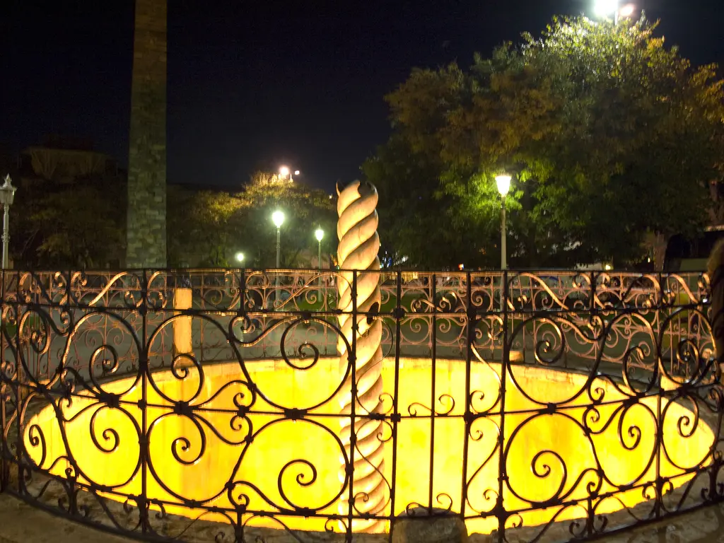 Gece Yılanlı Sütun; ışıkla aydınlatılmış sarı çukur, ortada tunç gövde, kıvrımlı parmaklık, arkada ağaçlar ve sokak lambaları.