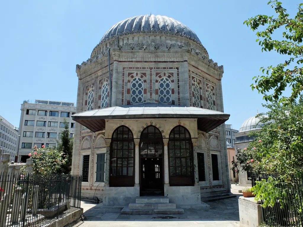 Şehzade Mehmed Türbesi dışarıdan; sekizgen gövde, dilimli kurşun kubbe, sütunlu giriş revağı, çini ve mermer kaplı cephe panoları ve önde güller.