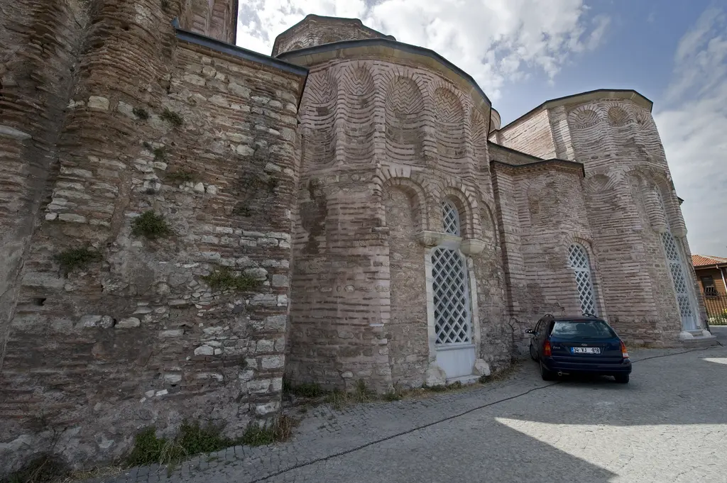 Zeyrek Camii'nin güneydoğu cephesi; kavisli apsis duvarında kırmızı tuğla ile açık renkli taş sıraları dönüşümlü uzanıyor, kemerli kafes pencereler ve önde park etmiş koyu renk otomobil.