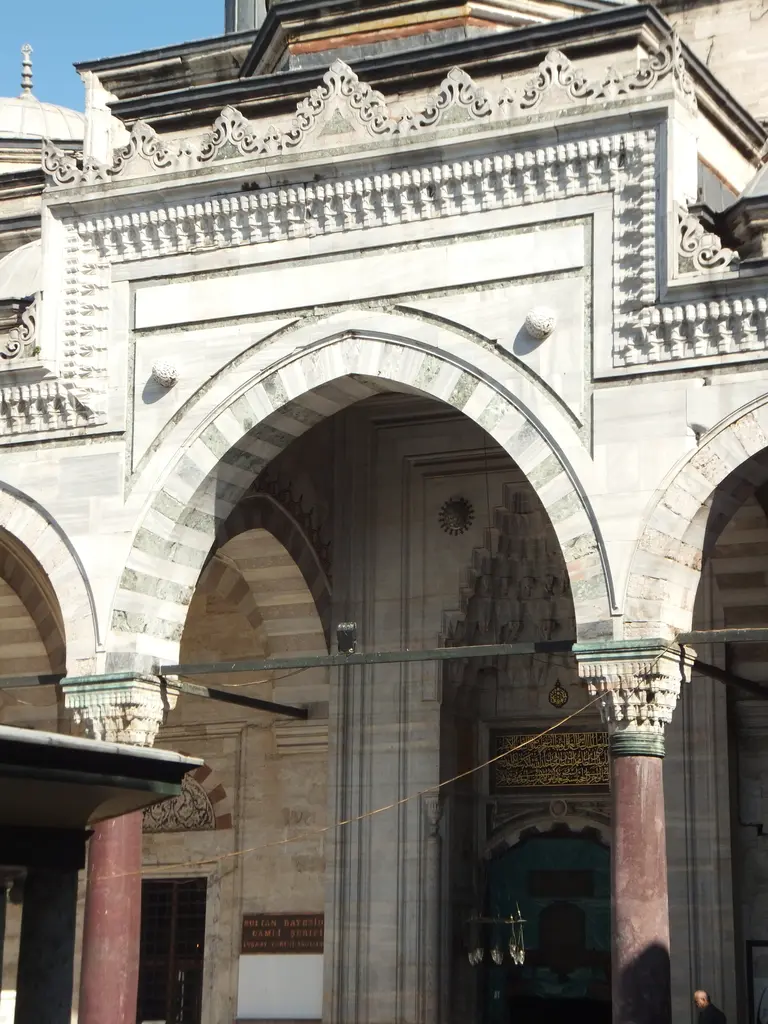 Avludan harime açılan taç kapı; mukarnaslı niş, iki yanda kırmızı sütunlar, üstte pencere sırası ve gölgeli kapı boşluğu.