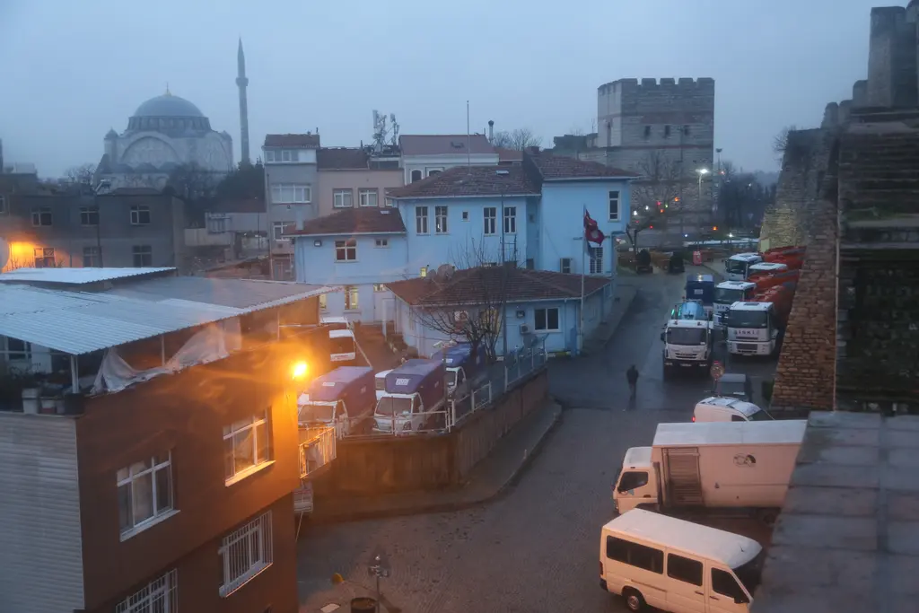 Surun üstünden mahalleye bakış; aşağıda dar sokak ve park etmiş minibüsler, evlerin ötesinde bir caminin kubbesi ve sur kuleleri.