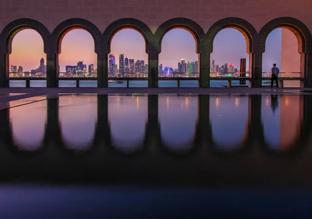 Doha'da kemerli bir galeri; sıra sıra yuvarlak kemerlerin arasından görünen gökdelenler ve önde suya vuran yansımaları.