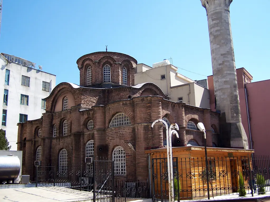Bodrum Camii'ne güneydoğudan bakış; sağda tek şerefeli kesme taş minare, ortada kubbe kasnağı, altta apsis çıkıntıları, önde sarı ahşap kulübe ve demir parmaklık.