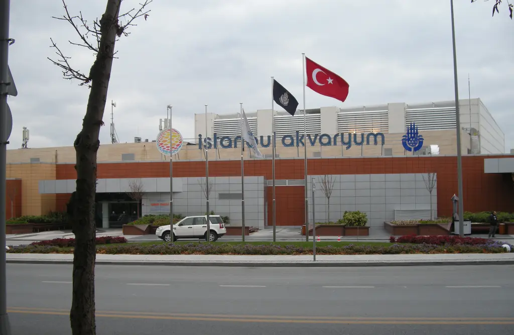 İstanbul Akvaryum'un caddeden görünen dış cephesi; turuncu ve bej panelli alçak yapı, çatı hizasında akvaryumun adı, bayraklar ve önde yol.