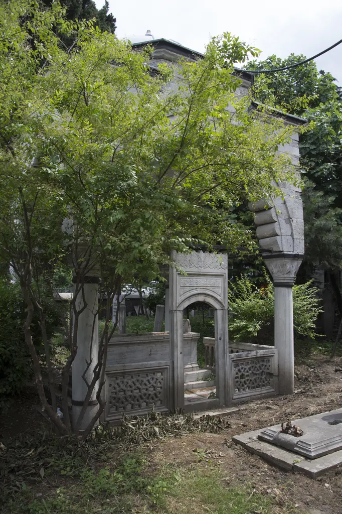 Defterdar Camii haziresinde açık türbe; ağaçların arasında dilimli kemerler, kabartmalı mermer korkuluk ve mezar taşları.