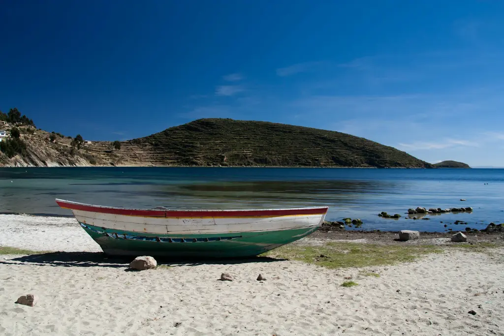 Bolivya'da Titicaca Gölü kıyısındaki Güneş Adası (Isla del Sol); kumsala çekilmiş kırmızı-beyaz-yeşil ahşap bir kayık ve karşıda teraslı tepe, derin mavi göl.