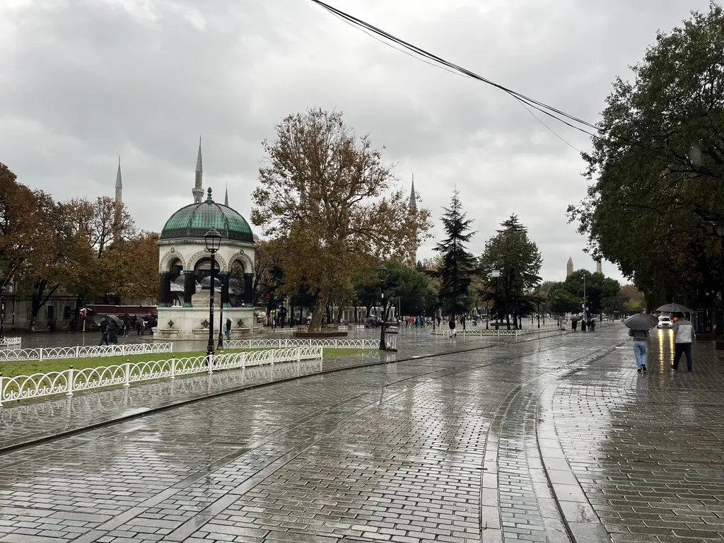 Yağmurlu bir günde Sultanahmet Meydanı; ıslak taş döşeli yol, beyaz demir parmaklıklar, ağaçlar ve yolun sonunda Alman Çeşmesi'nin kubbesi.