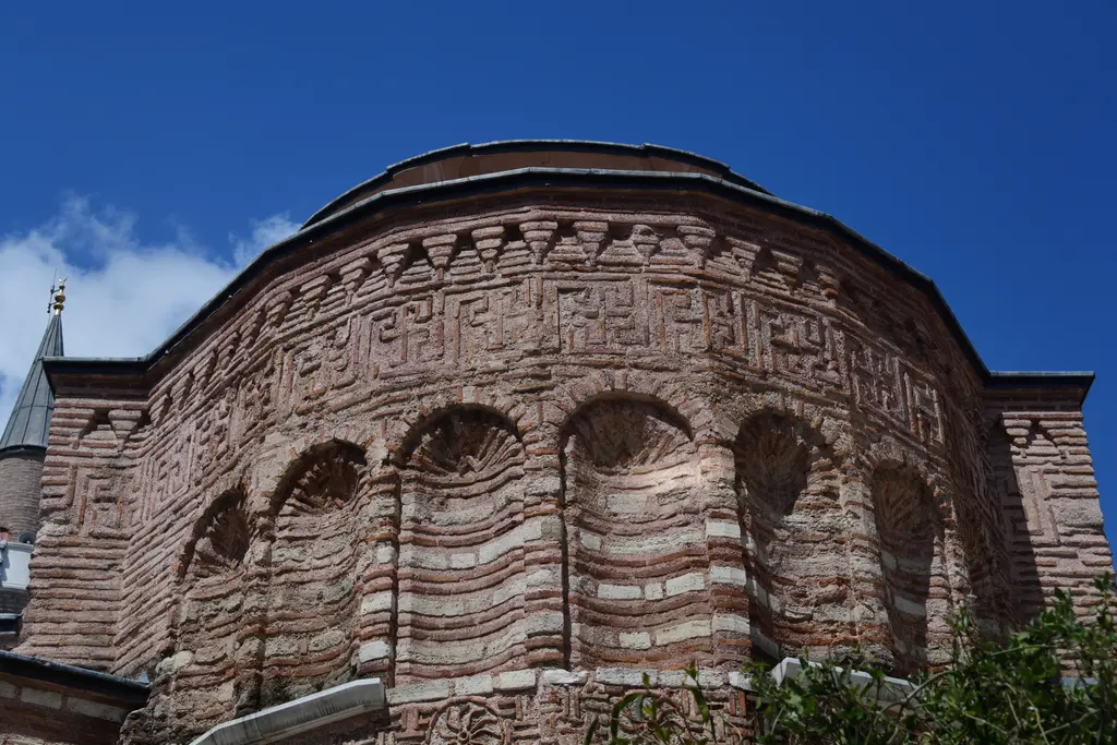 Fenari İsa Camii'nde apsisin kavisi ve saçak altındaki meander frizi; tuğlayla örülmüş kesintisiz anahtar motifi, altında kör kemer dizisi ve mavi gökyüzü.