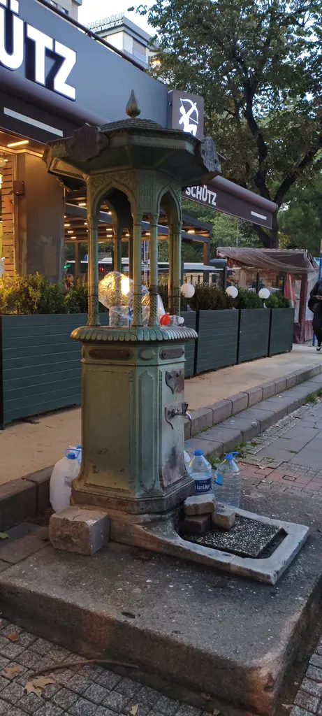 Göztepe'de Şehremaneti Çeşmesi; yeşil boyalı dökme demir sokak çeşmesi, musluklar ve arkasında kafe.