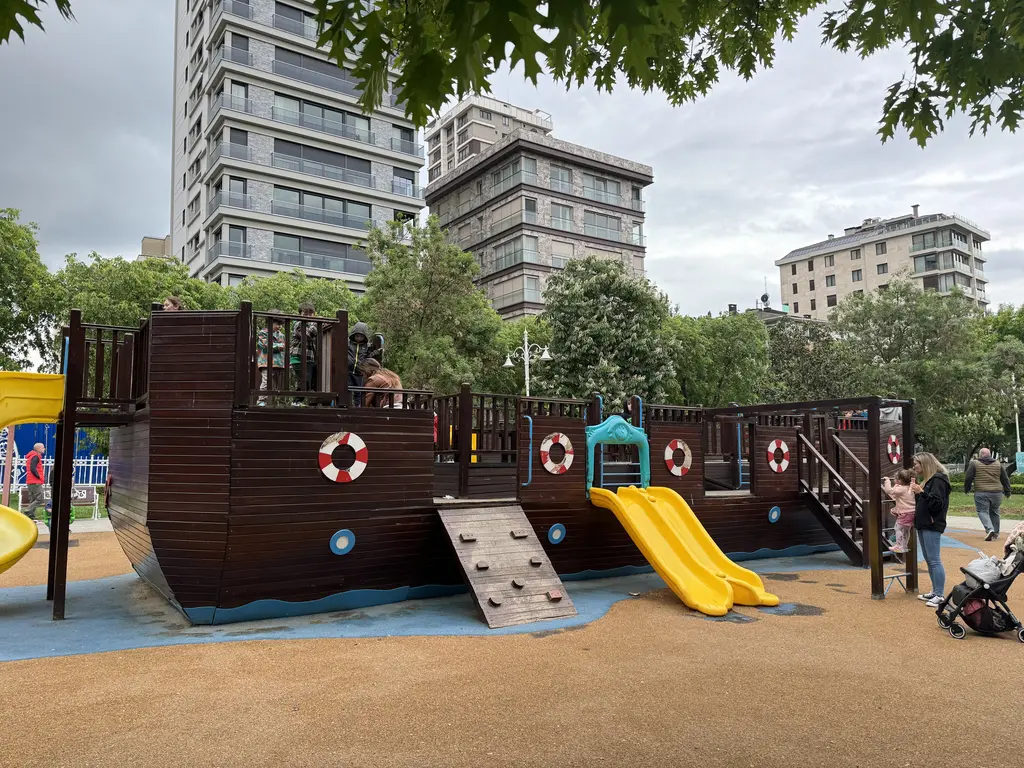 Göztepe Parkı'nda çocuk oyun alanı; ahşap gemi biçimli oyun yapısı, sarı kaydıraklar, ağaçlar ve arkada apartmanlar.