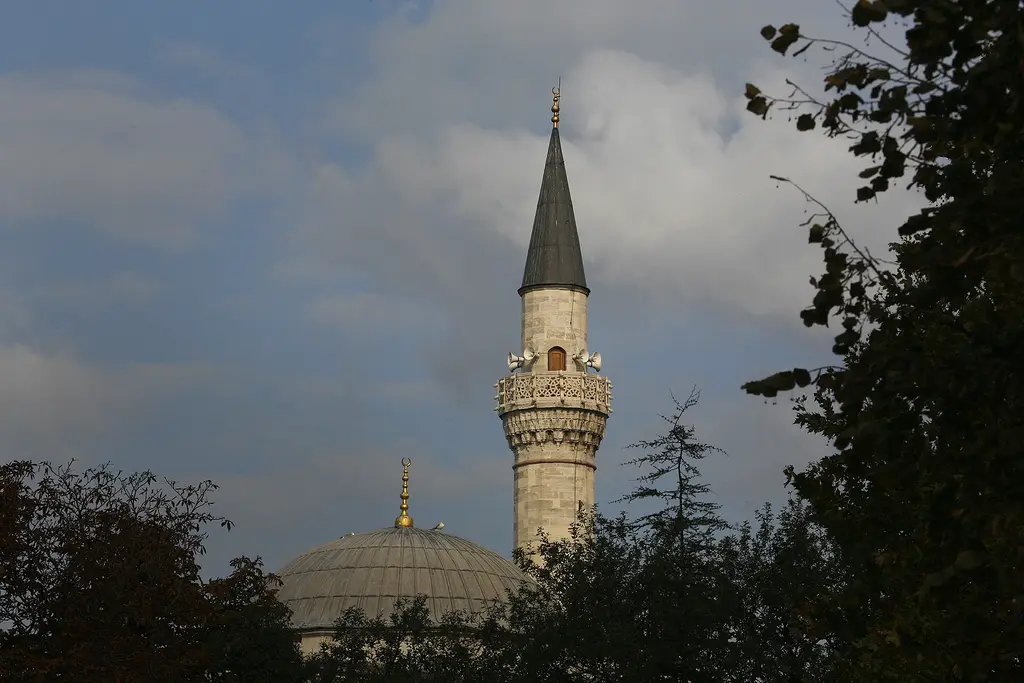 Firuz Ağa Camii'nin minaresi ve kubbesi; mukarnaslı şerefe, sivri külah, ağaçların arasından görünen kurşun kubbe ve bulutlu gök.