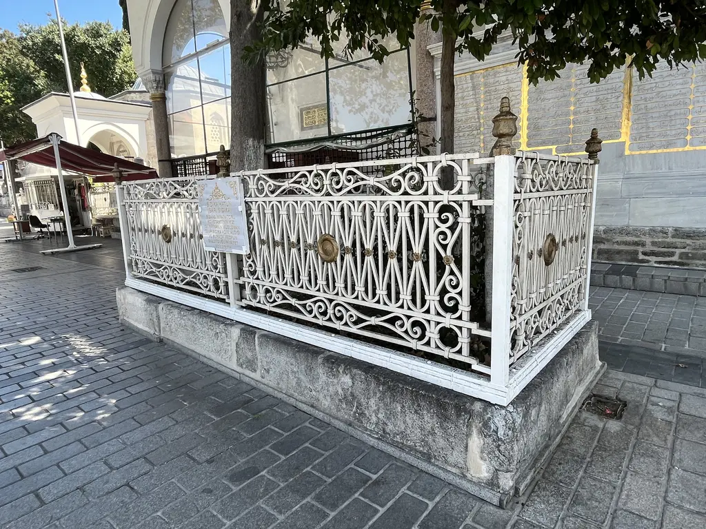 Koca Mustafa Paşa Camii'nin önünde taş kaide üstünde açık bir kabir; beyaz işlemeli dökme demir parmaklık, altın madalyonlar, küçük mermer levha ve arkada caminin camlı revağı.