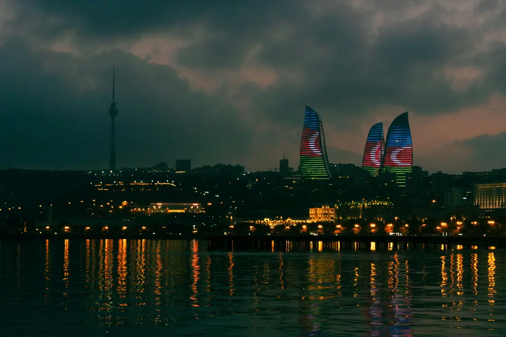 Bakü'nün gece silueti; körfezin karşısında ışıklı kule sıraları, üzerinde Azerbaycan bayrağı beliren Alev Kuleleri ve suda uzayan yansımalar.