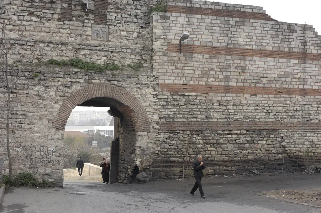Yedikule Kapısı; sur duvarını delen tuğla kemerli geçit, duvarda taş ile tuğlanın almaşık bantları, kemerin altından görünen şehir ve önde yürüyen birkaç kişi.