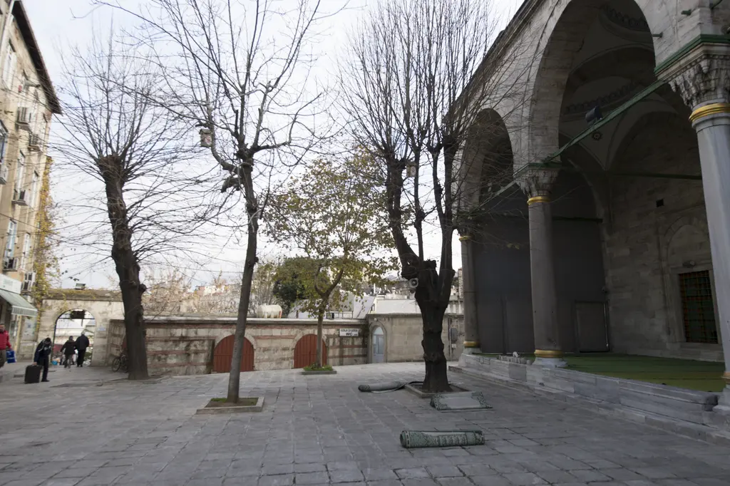 Gazi Atik Ali Paşa Camii'nin avlusu; taş döşeme, yapraksız ağaçlar, alçak avlu duvarı, sağda revak kemerleri ve uzakta yürüyen insanlar.
