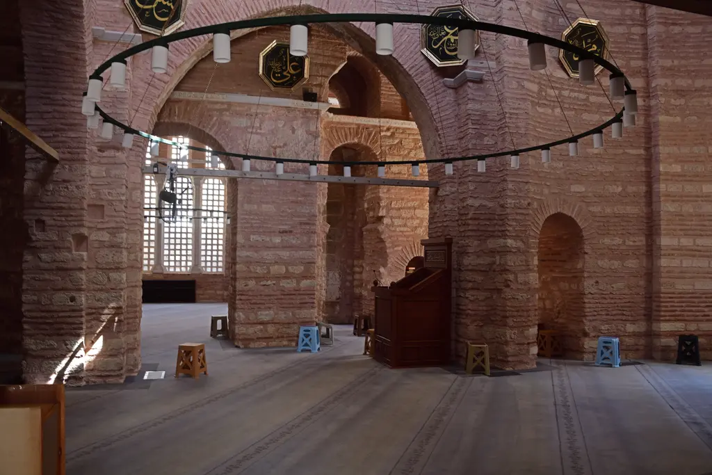 Fenari İsa Camii'nin harimi; sıvası sökülmüş tuğla taş payeler ile kemerler, tavandan sarkan siyah demir avize çemberi, sağda ahşap kürsü ve gri saflı halı.