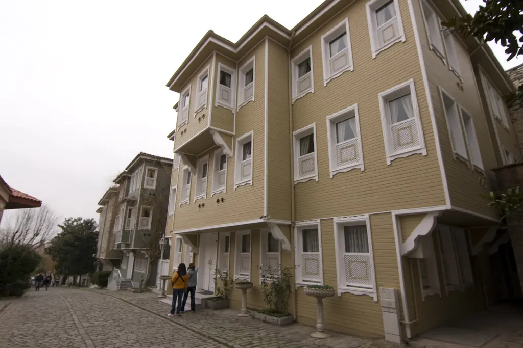 Soğukçeşme Sokağı'nda yan yana sıralanan sarı ahşap evler; beyaz pencere çerçeveleri, çıkma katlar, önde yürüyenler ve kapalı gök.