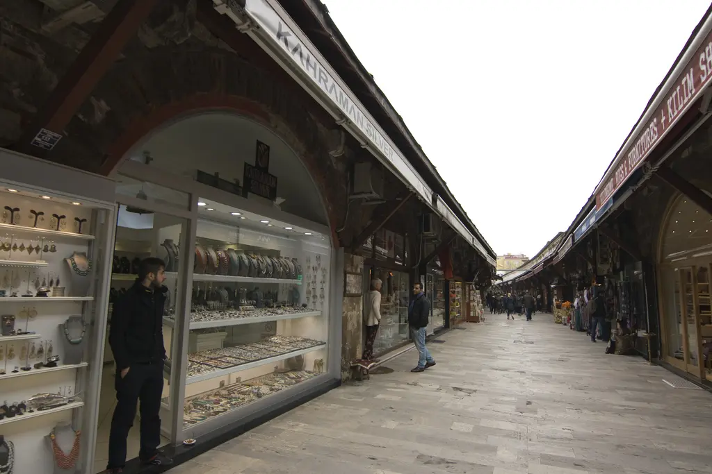 Arasta Çarşısı'nda cam vitrinli dükkânlar; raflarda seramik ve hediyelik eşyalar, önde duran bir esnaf, arkada uzanan kemerli sıra.