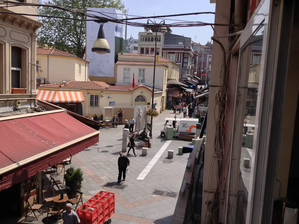 Kadıköy Çarşısı'nda sokak; tenteli dükkânlar, sokak lambası, yürüyen insanlar ve ileride kilise çatısı.