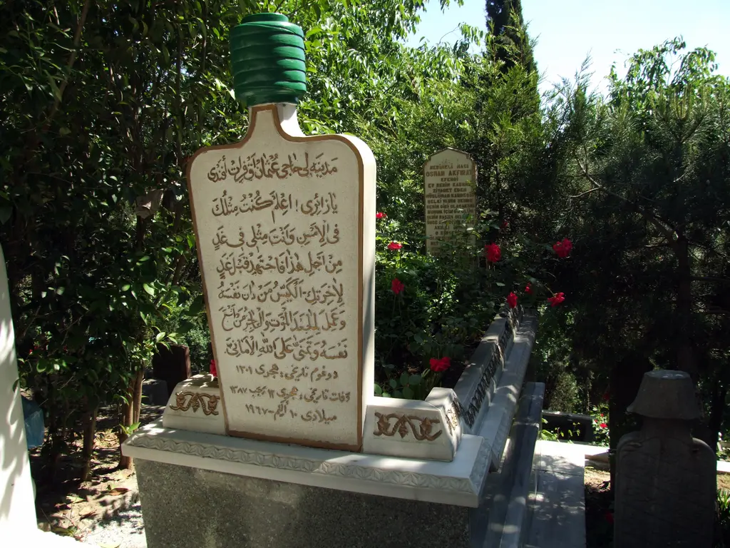 Eyüp Mezarlığı'nda mezar taşı; yeşil başlıklı beyaz taşta yaldızlı Arap harfli yazı, çevresinde güller ve ağaçlar.