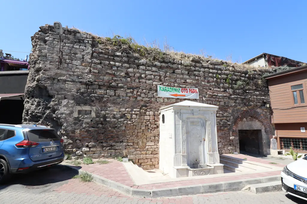 Cumhuriyet Çeşmesi; eski taş duvarın önünde beyaz mermer çeşme, yanında park etmiş araba ve ahşap kaplı ev.