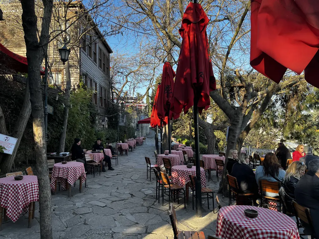 Pierre Loti Tepesi'nde çay bahçesi; taş döşeli yol, kırmızı şemsiyeler, damalı örtülü masalar, ağaçlar ve oturan ziyaretçiler.