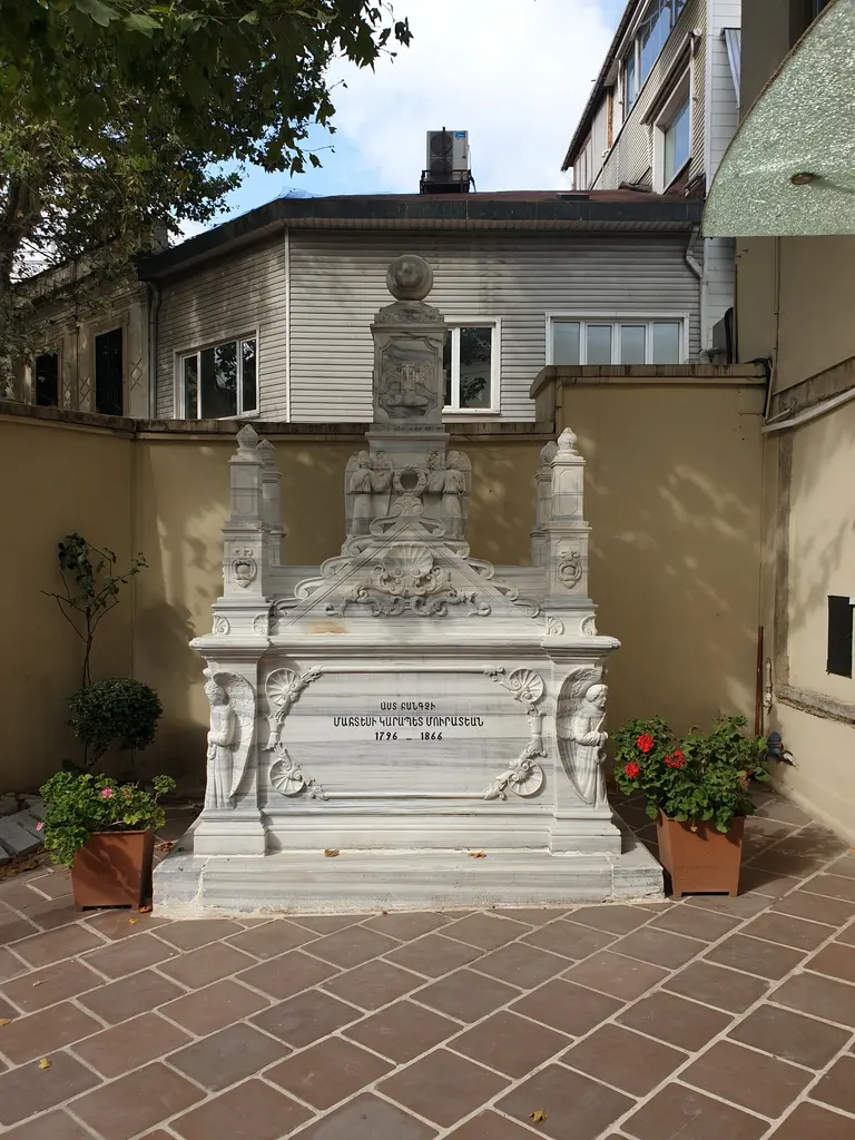Surp Takavor Kilisesi'nin avlusunda kabir; süslü mermer lahit, Ermenice kitabe, saksılarda çiçekler ve arkada evler.