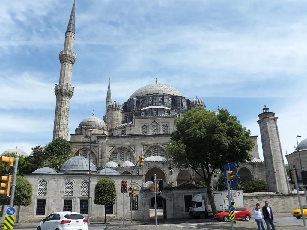 Şehzade Camii üç çeyrek açıdan; avlu duvarının üstünde yükselen kubbe pramidi, iki bezemeli minare, önde bir ağaç ile trafik ışıkları ve mavi gökyüzü.