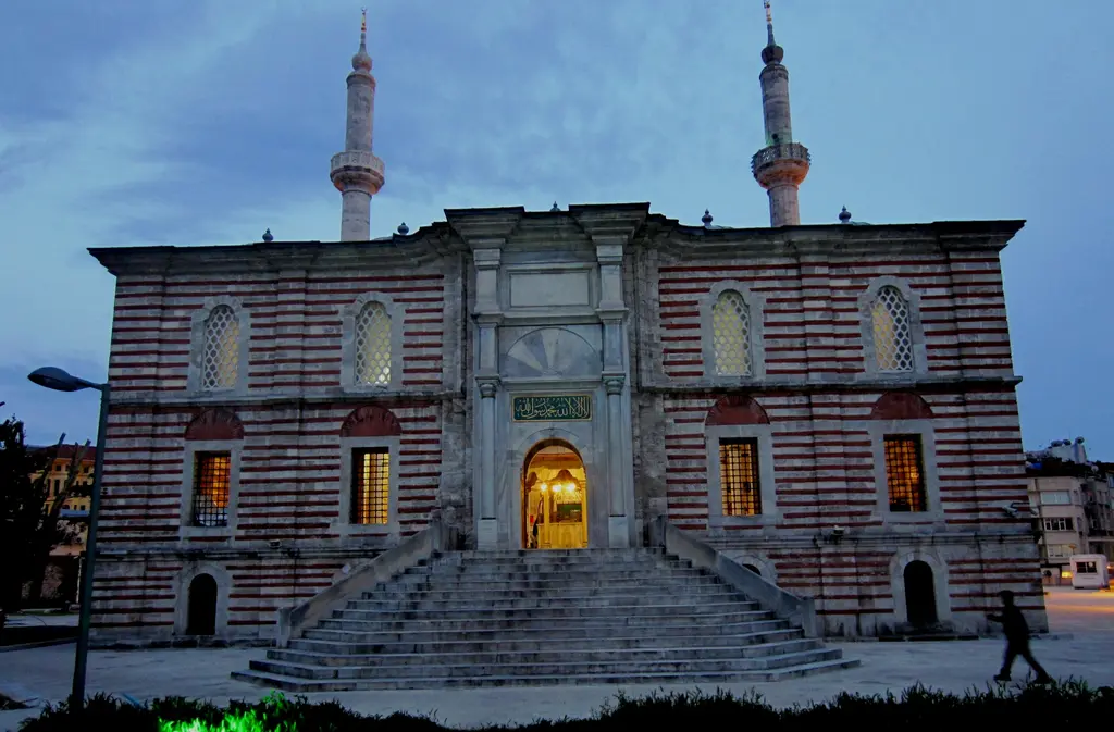 Akşam saatinde Laleli Camii; aydınlatılmış şeritli cephe, iki minare, önünde geniş merdiven ve caddeden geçen bir yaya.
