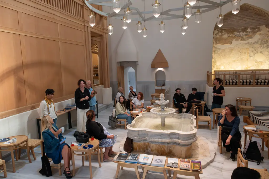 Çinili Hamam'ın soğukluğu; ortada mermer şadırvan, çevresinde ahşap sedirler ve alçak sehpalar, tavanda halka avize, oturan kalabalık.