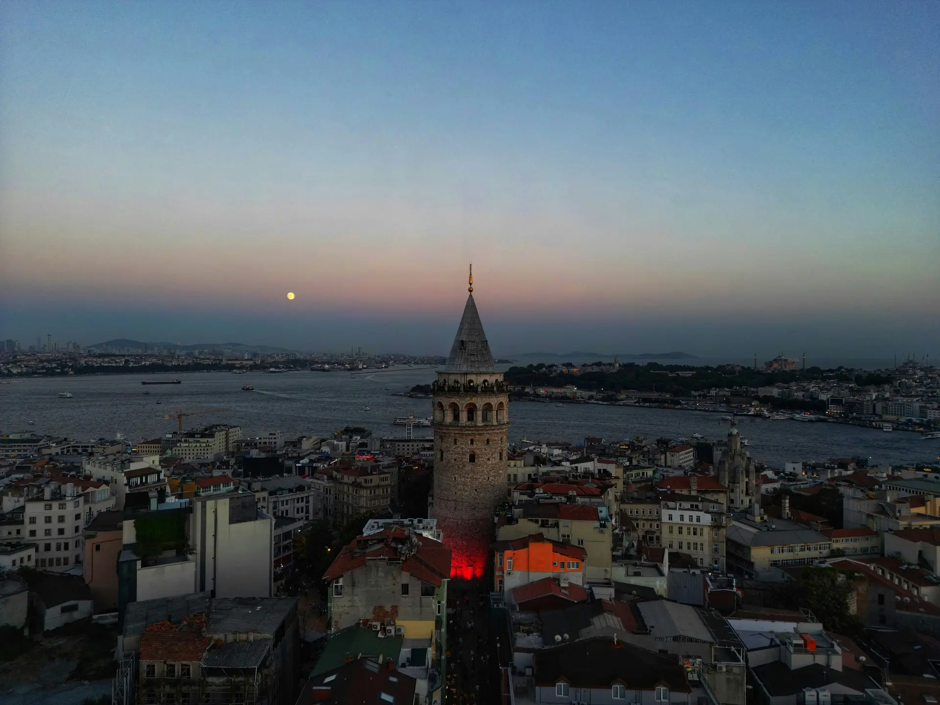Havadan Galata Kulesi alacakaranlıkta; önde kiremitli çatılar ve kuleye uzanan dar sokak, arkada Haliç'in ağzı, tarihi yarımada ve ufukta dolunay.