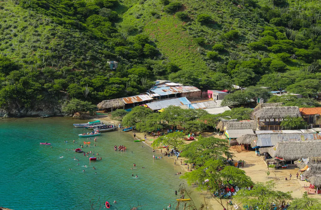 Santa Marta yakınında koy; turkuaz sığ suda yüzen kalabalık, kumsala dizilmiş sazdan çatılı barakalar ve arkadaki yeşil tepeler.