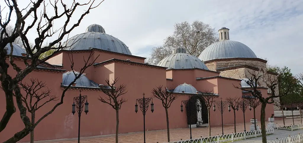 Sultanahmet'te Haseki Hürrem Sultan Hamamı; somon rengi sıvalı uzun duvar, arkasında sıralanan kurşun kaplı kubbeler ve önünde çıplak ağaçlarla demir korkuluk.