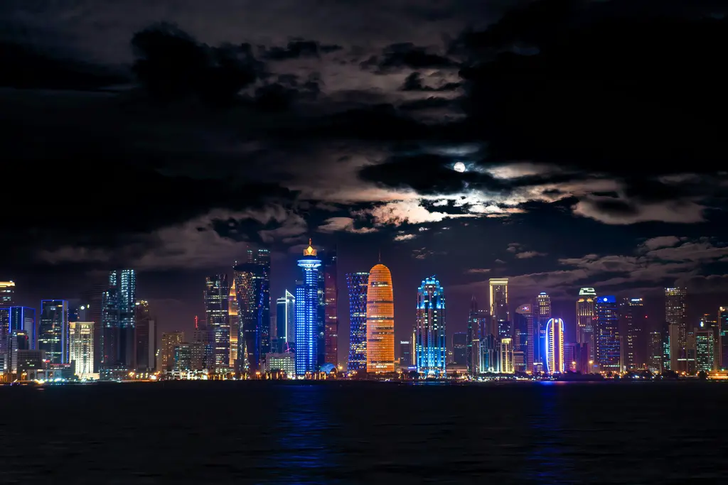 Doha'da gece silueti; ışıklandırılmış gökdelen sırası, üzerinde beliren dolunay ve suya vuran renkli yansımalar.