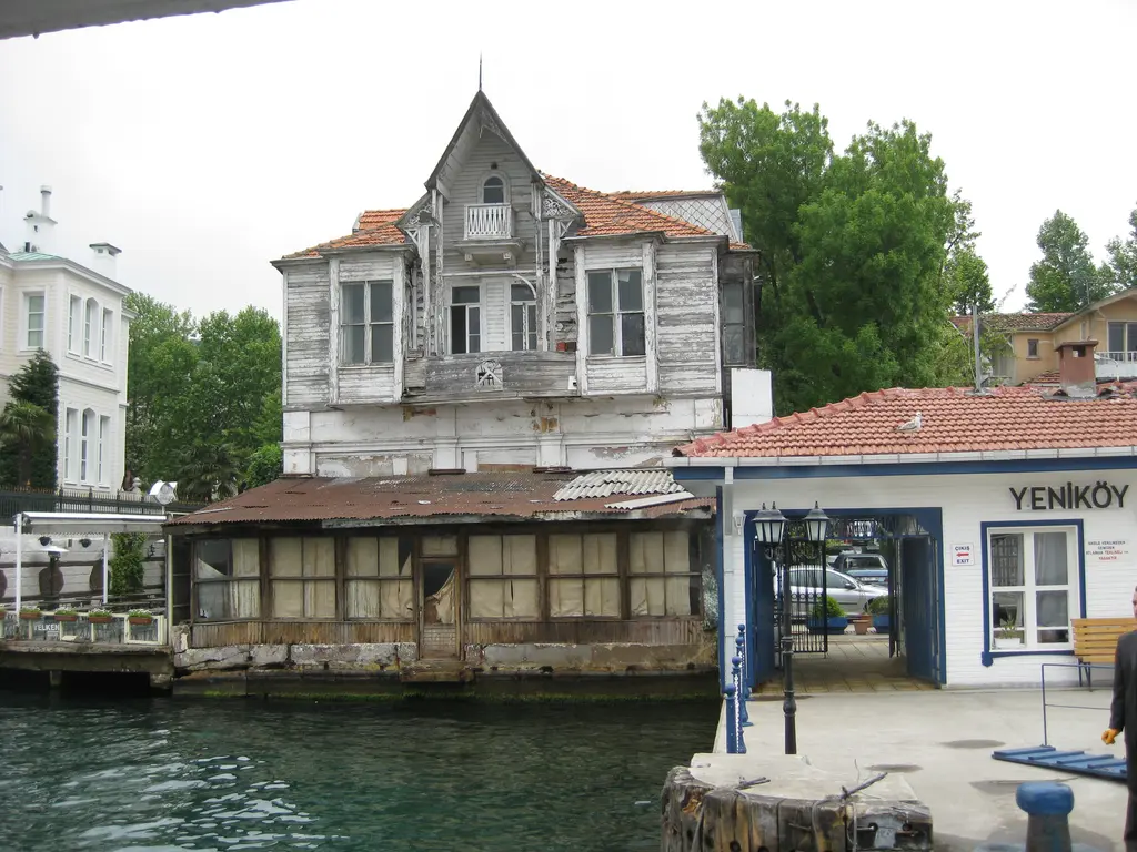 Yeniköy İskelesi; mavi beyaz iskele binası, Yeniköy tabelası, yanında boyasız ahşap ev ve arkada yeşil ağaçlar.