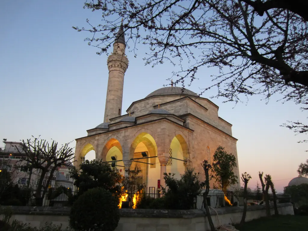 Akşam ışığında Firuz Ağa Camii; aydınlatılmış kemerli revak, sarı ışık alan taş duvarlar, kubbe, minare ve önde budanmış ağaçlar.