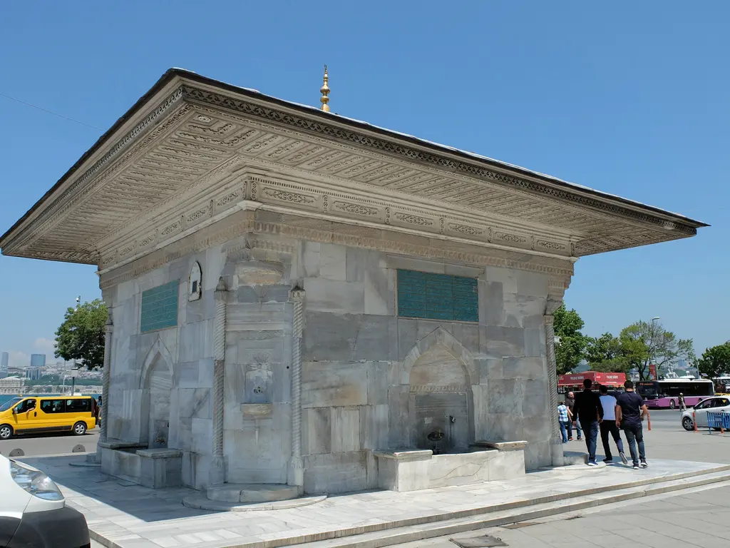 III. Ahmed Çeşmesi; köşeden görünen mermer gövde, saçak, kitabe levhaları ve önünde duran insanlar.