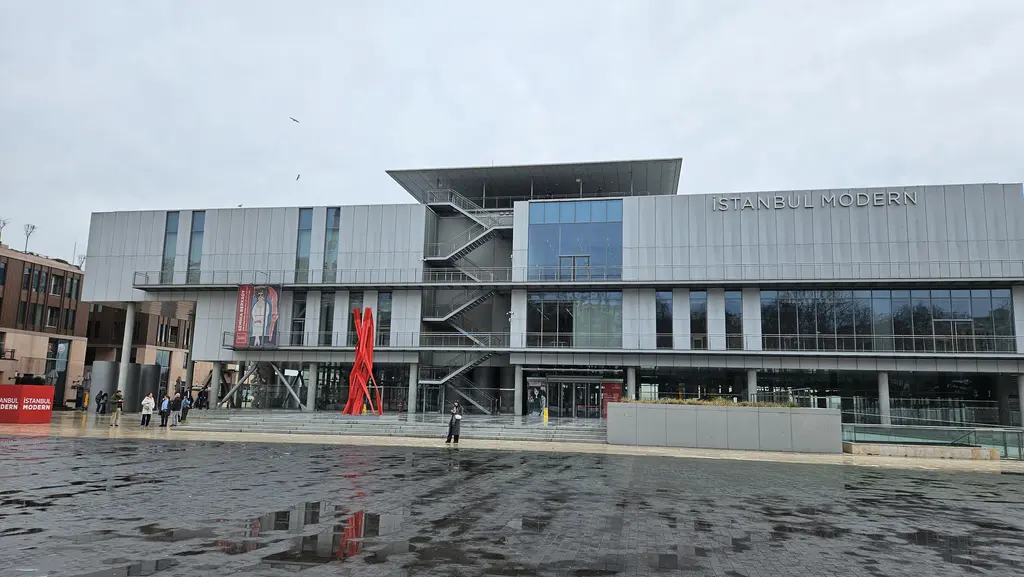 Kapalı havada İstanbul Modern'in cephesi karşıdan; ıslak taş meydan, kırmızı bir heykel ve girişteki ziyaretçiler.