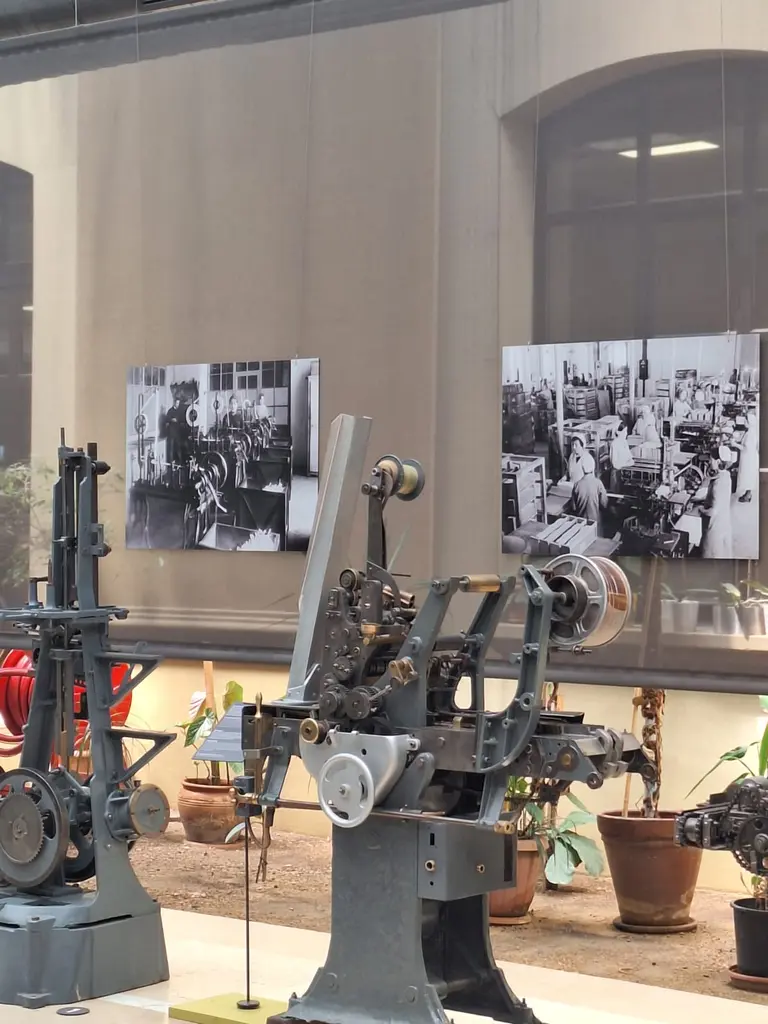 Tütün işleme makineleri; metal gövdeli iki makine, arkada fabrikada çalışan kadınların siyah beyaz fotoğrafları ve saksılar.