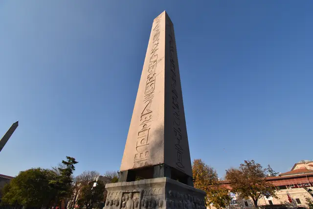 Sultanahmet Meydanı'nda Dikilitaş'a alttan bakış; pembe granit gövde, hiyeroglif sütunu, mermer kaide, sonbahar ağaçları ve koyu mavi gökyüzü.