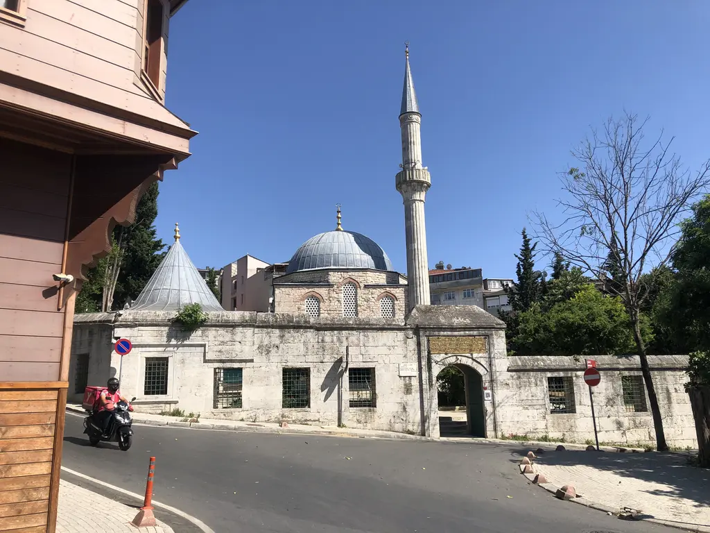 Çinili Külliyesi; cadde kenarında uzun taş avlu duvarı, şadırvan çatısı, kubbe, minare ve geçen bir motosiklet.