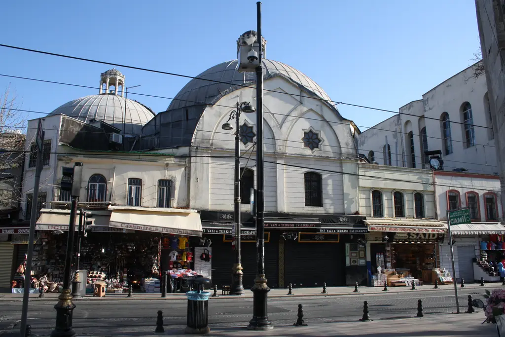 Çemberlitaş Hamamı'na sokaktan bakış; kurşun kaplı büyük ve küçük iki kubbe, kemerli alınlık, altta sıra dükkânlar ve önde sokak lambaları.