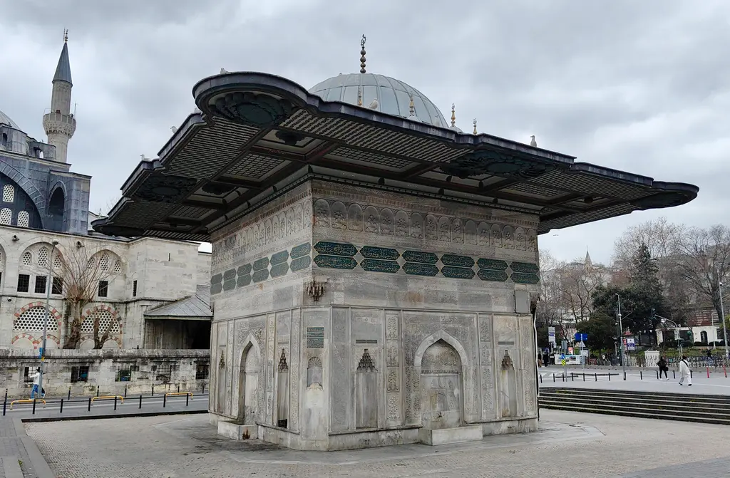 Tophane Çeşmesi ve arkasında Kılıç Ali Paşa Camii; bulutlu gökyüzü, geniş saçak, kubbe ve minare, önde taş döşeli meydan.