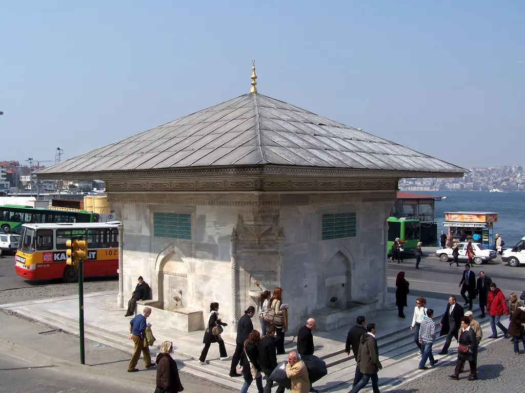 Üsküdar III. Ahmed Çeşmesi; mermer çeşme, önünde yürüyen kalabalık, arkada otobüsler, vapur ve deniz.