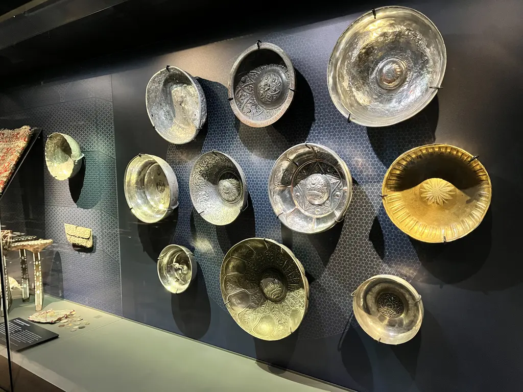 Çinili Hamam Müzesi'nde hamam tasları; koyu duvara asılı on kadar kabartma bezemeli pirinç ve gümüş tas, yanda küçük eşya vitrini.