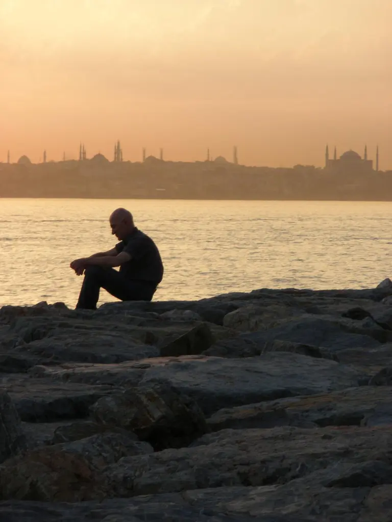 Moda sahilinde gün batımı; kayaların üstünde oturan bir adamın silueti, turuncu gökyüzü ve ufukta tarihi yarımada.