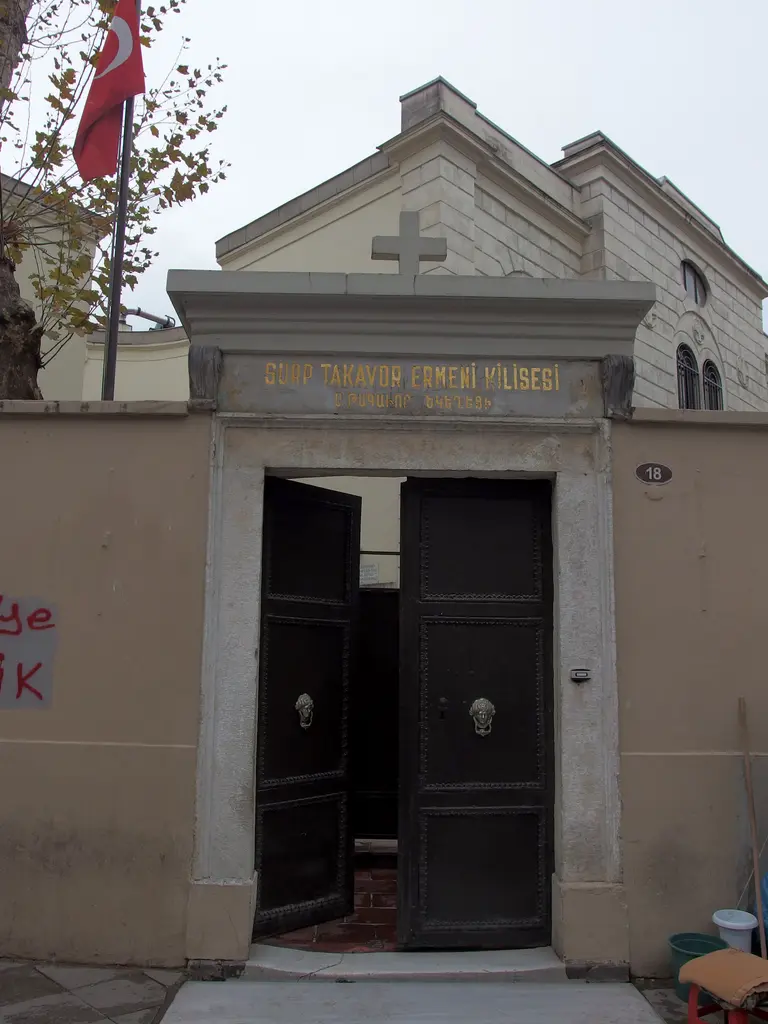 Surp Takavor Kilisesi'nin girişi; haçlı alınlık, açık ahşap kapı, beyaz taş çerçeve ve yanında bayrak.