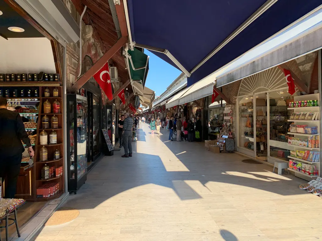 Arasta Çarşısı'nda güneşli bir gün; iki yanda dükkânlar, lacivert tenteler, asılı Türk bayrakları ve sokakta yürüyen ziyaretçiler.